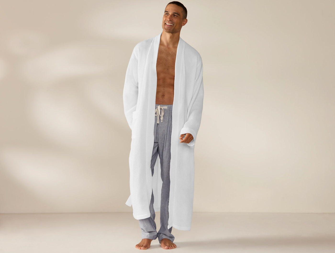 Man wearing open Coyuchi white waffle robe over grey striped pants, full-length front view.