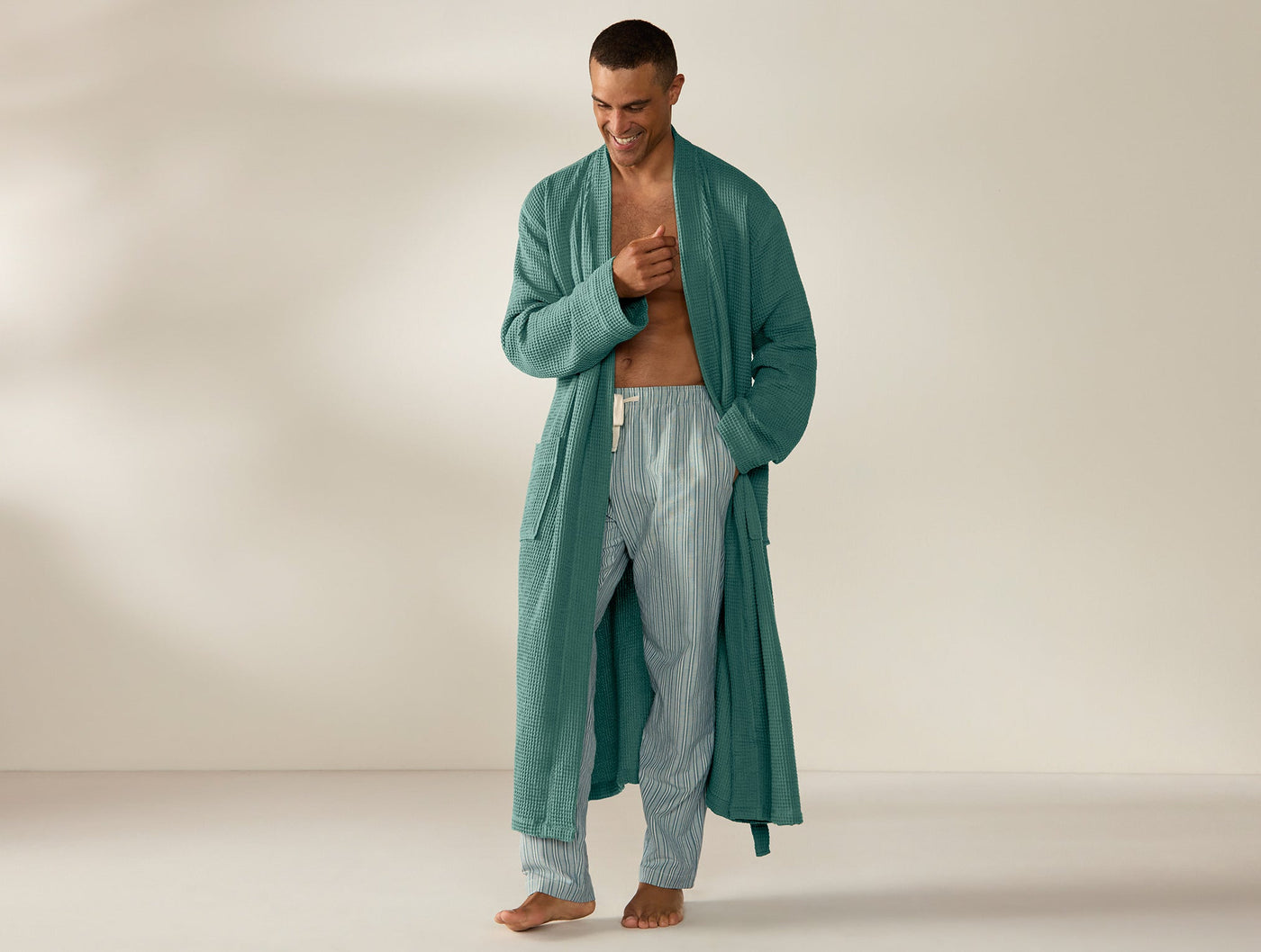 Man wearing Coyuchi long waffle robe in teal, open over striped pajama pants.