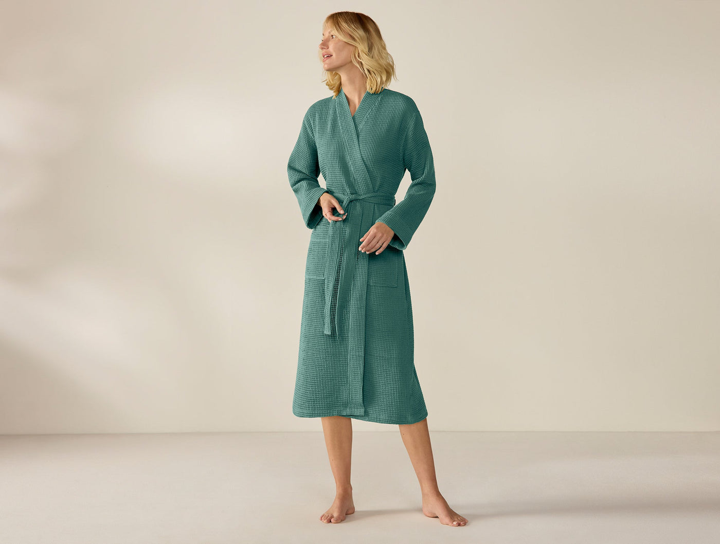 Model wearing Coyuchi teal waffle-knit unisex robe, belted at waist, midi length.