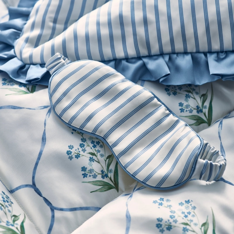 Blue and white striped silk sleep mask resting on floral-printed bedding with ruffled blue trim detail.