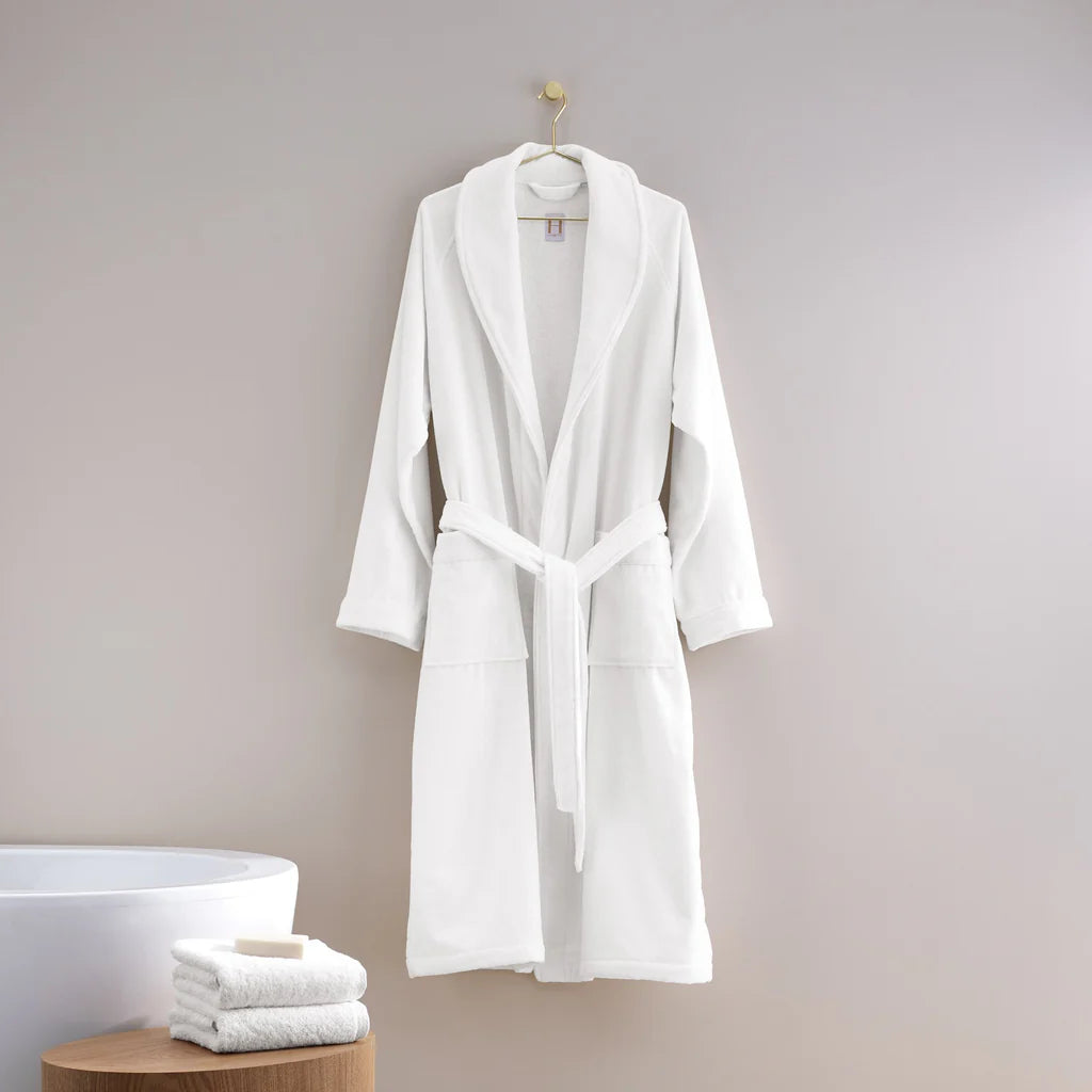 The Shawl Collar Robe by Frette hangs on a hook against a light gray wall, with folded white towels and a bar of soap arranged on a wooden stool nearby.