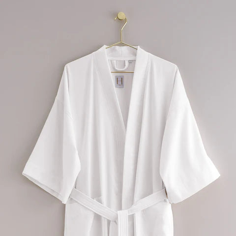 The Pique Kimono by Frette, a white lightweight robe with kimono sleeves, hangs on a gold hook against a light grey wall.