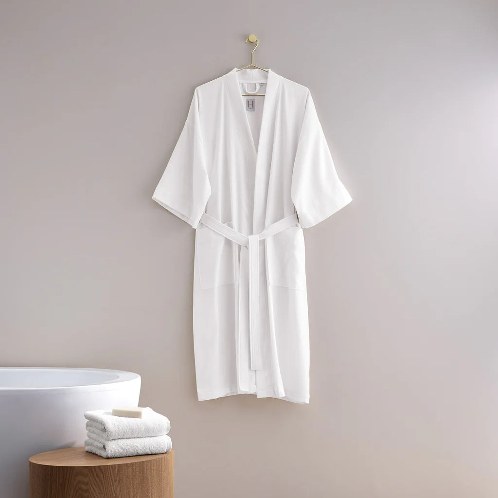 The Pique Kimono bathrobe by Frette hangs on a hook against a light gray wall, with a stack of folded white towels on a wooden stool next to the bathtub.