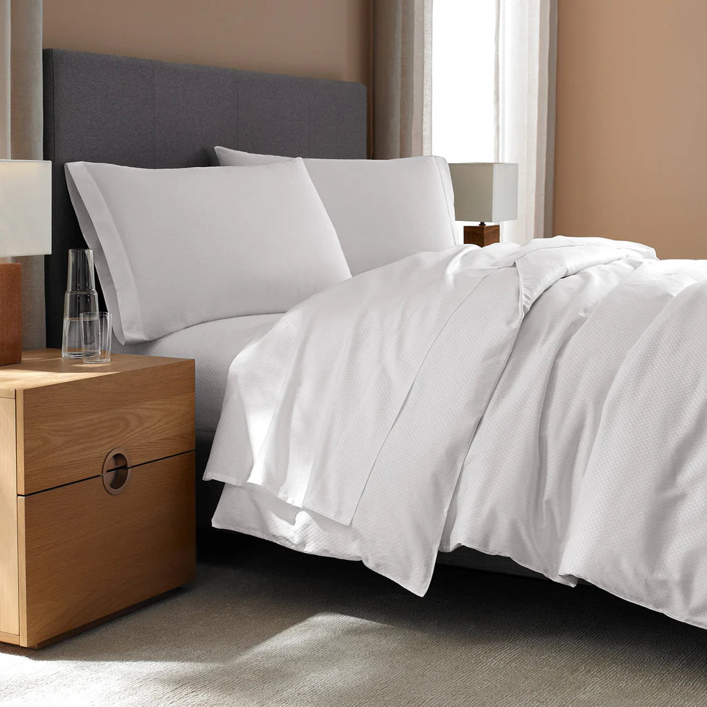 A modern, neutral-toned bedroom features a neatly made bed with Frette’s Checkered Sateen Pillow Sham Set, silky white bedding, and a wooden nightstand holding two glasses and a carafe.