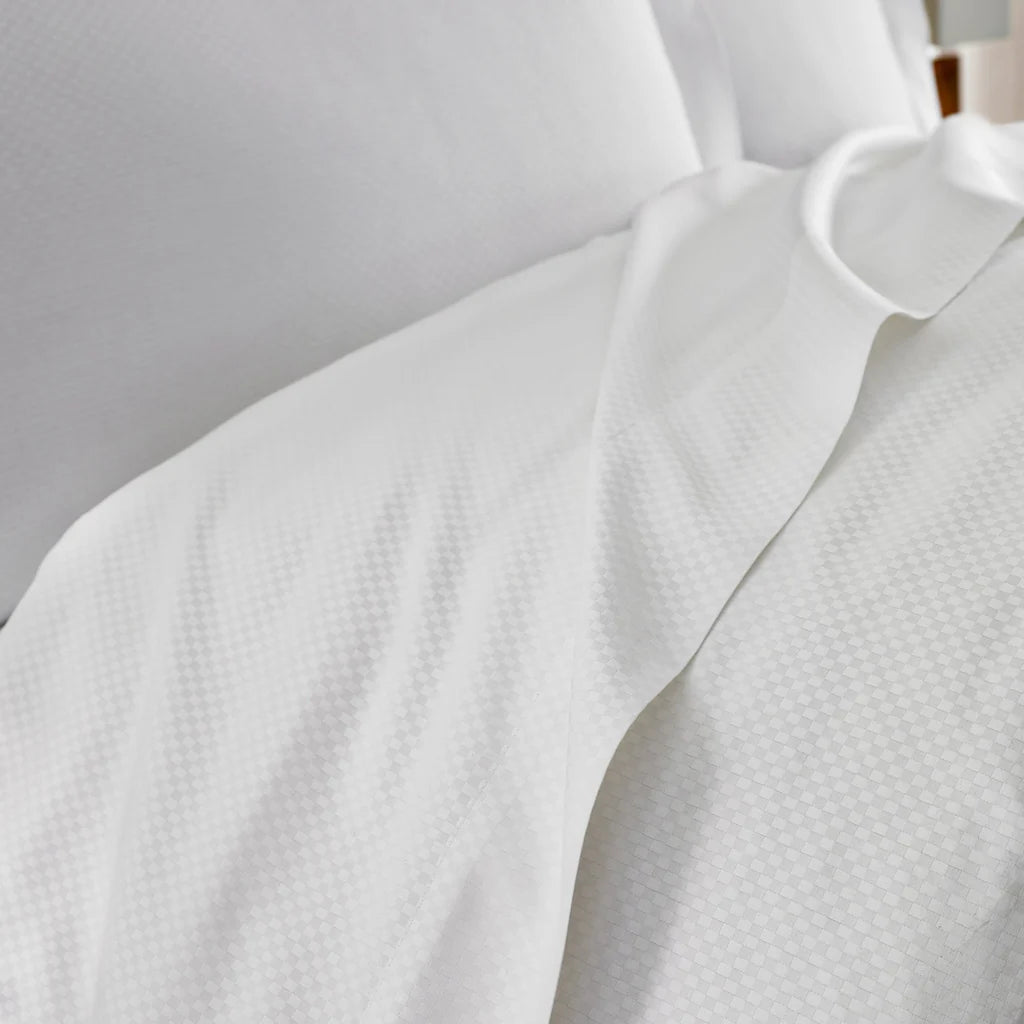 Close-up of a neatly made bed featuring Frette's Checkered Sateen Pillow Sham Set, showcasing white textured bedding and pillows for a touch of silky luxury.