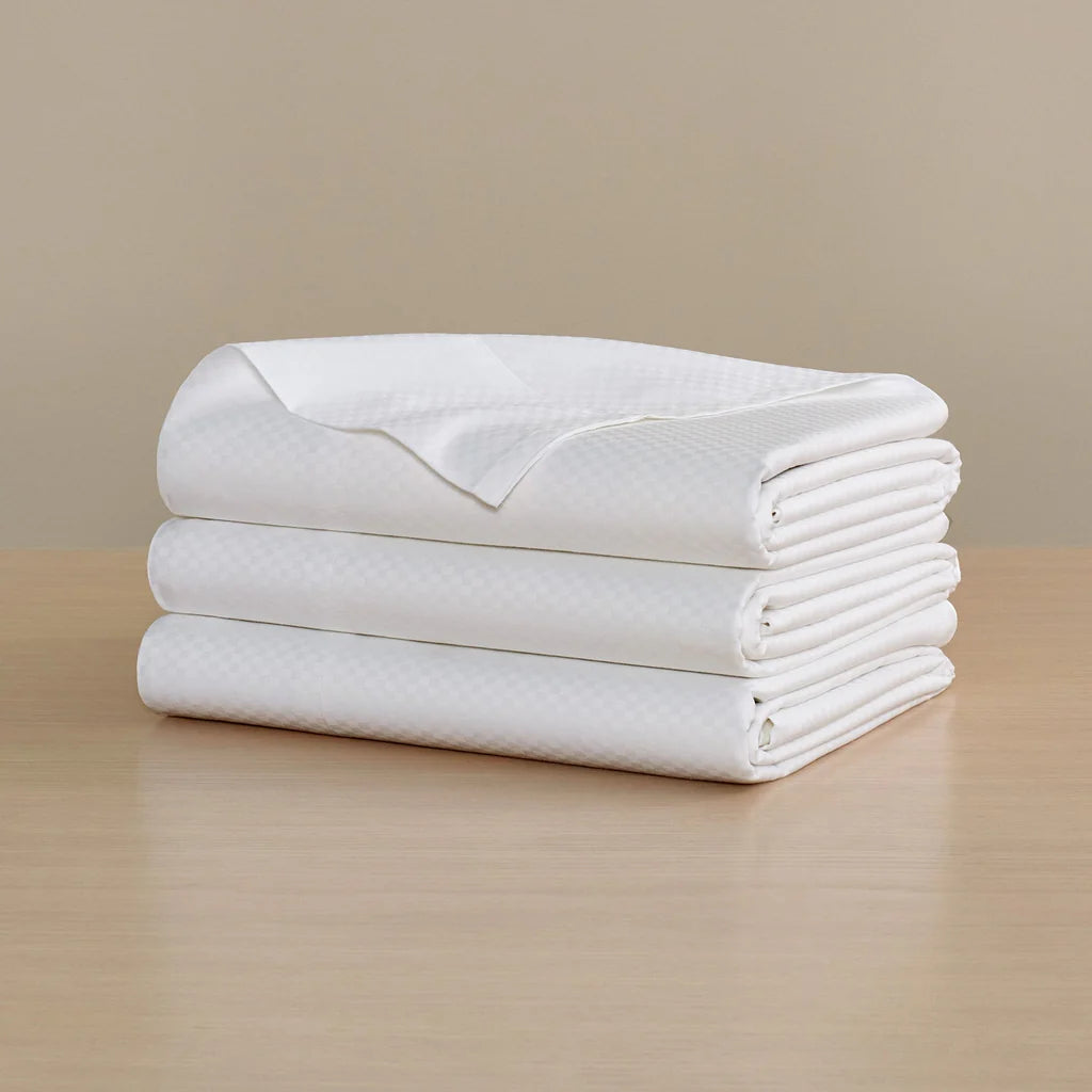 The Frette Checkered Sateen Flat Sheet, featuring a subtle checkered pattern in white, is neatly folded on a light wood surface against a beige background.