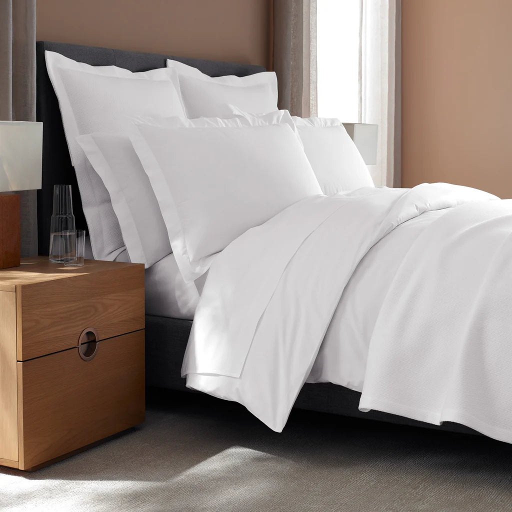 A neatly made bed with white bedding and Frette's Waves | Pique Euro Sham Set sits beside a wooden nightstand with a glass carafe and glass, evoking a sense of hotel hospitality.