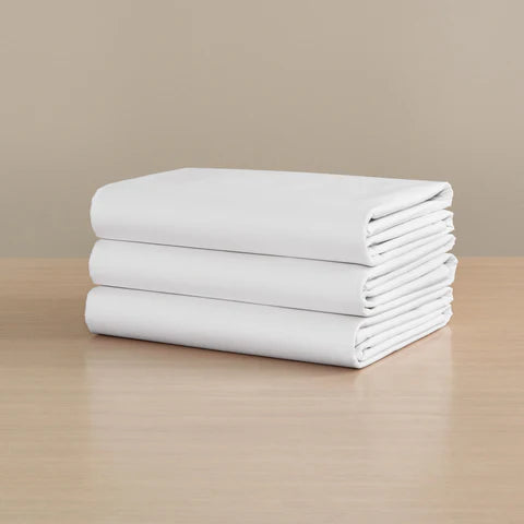 Three Frette Percale Fitted Sheets by Frette, crafted from long-staple cotton, are neatly folded and stacked on a light wooden surface against a beige background.