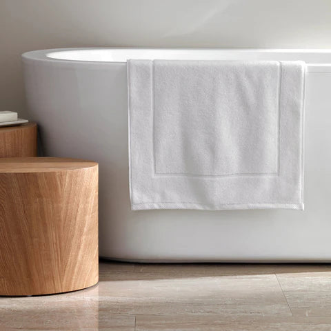 The Frette Simple Border Bath Mat, in white 100% cotton, rests by a modern tub beside a wooden stool on a light-tiled floor.