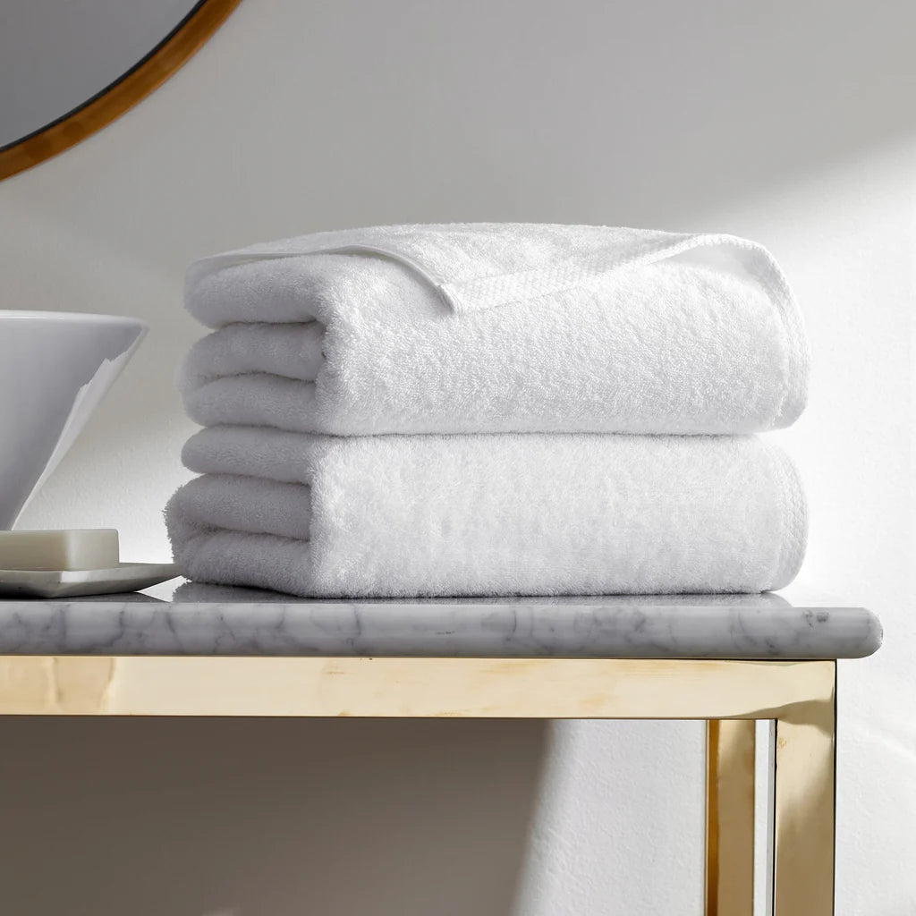 A stack of three Frette Simple Border Bath Sheet Set towels sits neatly folded on a marble countertop with a gold metal frame, beside a white sink and a round mirror.