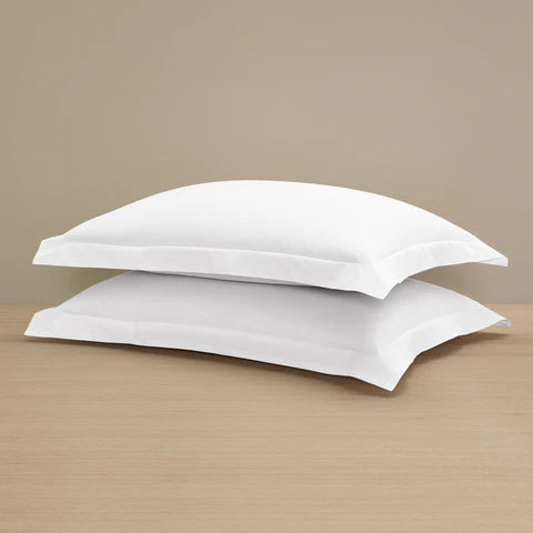 Two Frette Sateen Pillow Shams by Frette, crafted from luxurious long-staple cotton, are stacked on a light wooden surface against a plain beige background.