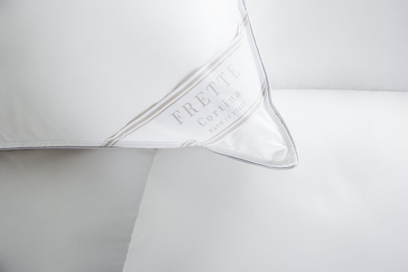 Close-up of the Frette Cortina | Down Pillow in white cotton sateen, featuring a label that reads "Frette Cortina Made in Italy," displayed on a white bed.