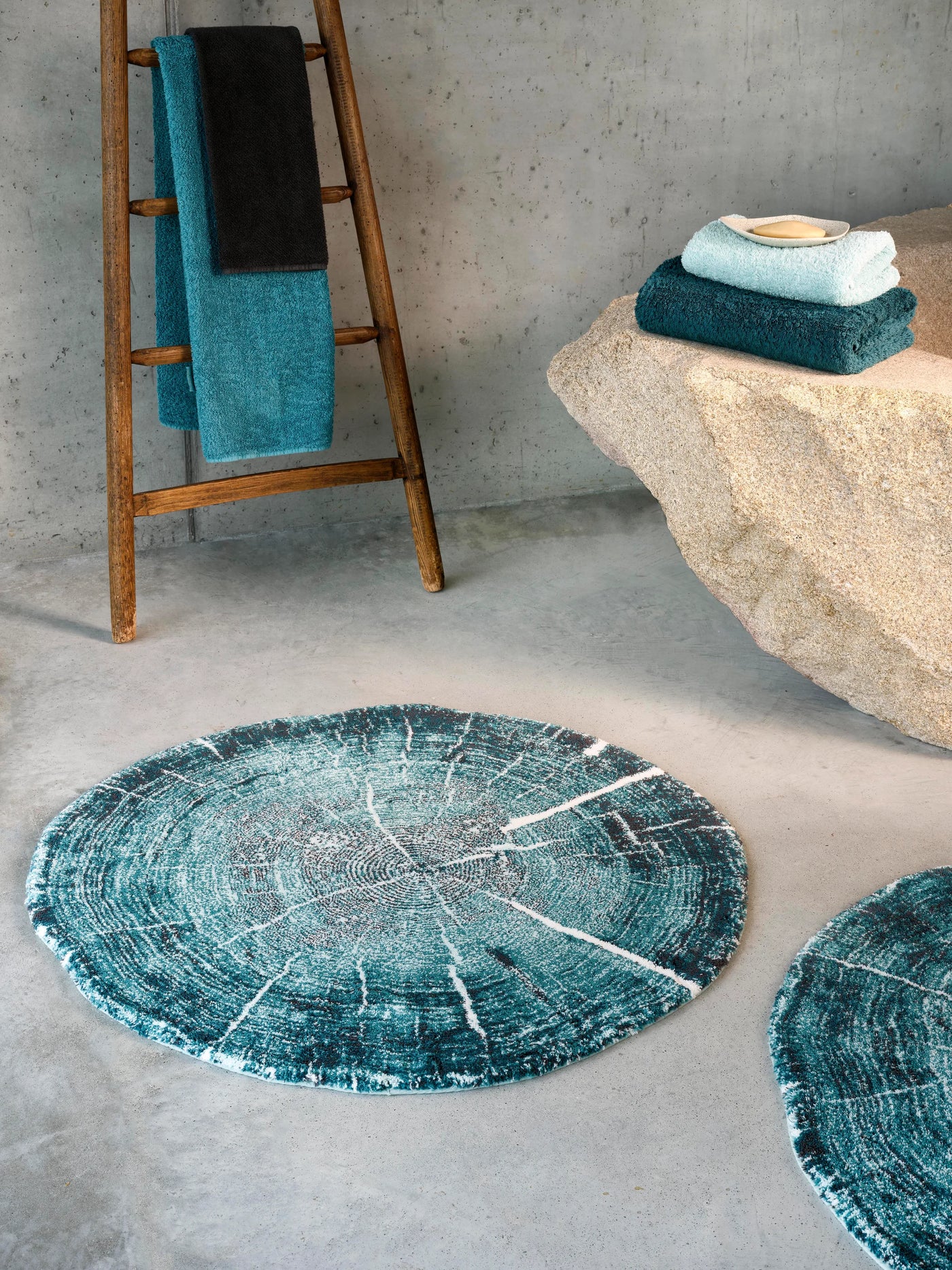 Teal bath rug with tree ring pattern, styled on concrete floor beside stone and wooden ladder.