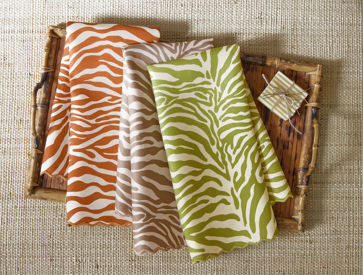 Three Regine printed linen guest towels by Matouk in orange, beige, and green are folded on a bamboo tray next to a small wrapped gift, all arranged on a textured beige surface.