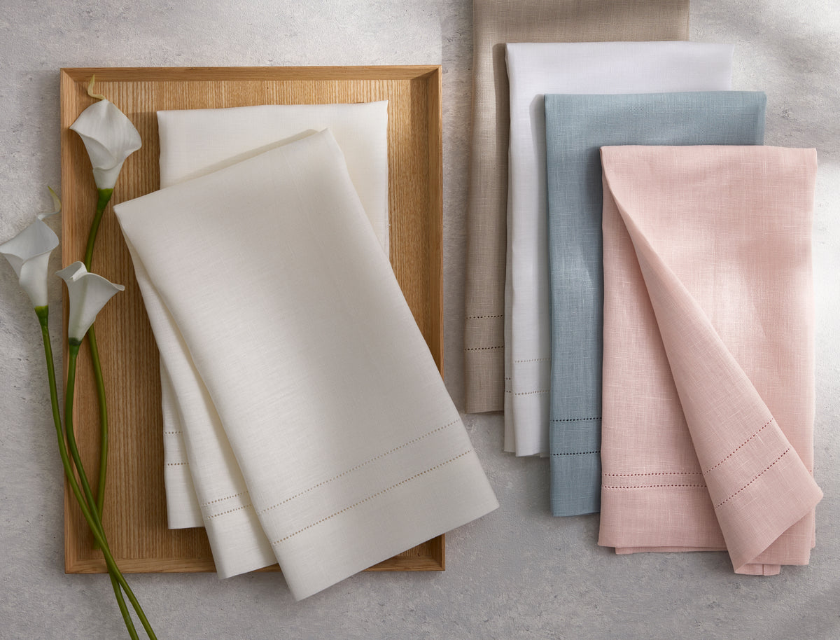 The Valentina Hemstitch Napkin Set by Matouk includes four folded linen napkins in white, beige, blue, and pink, elegantly displayed on a wooden tray with calla lilies—an ideal choice for an elegant tabletop.