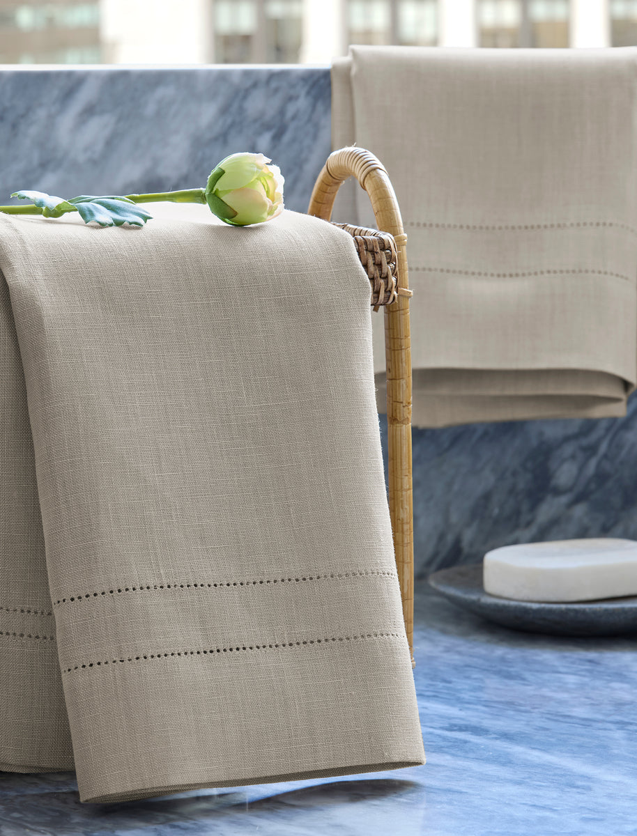 The Valentina Hemstitch Guest Towel by Matouk in beige linen, elegantly draped over a wicker basket and topped with a green and white flower, rests on a blue marble surface.