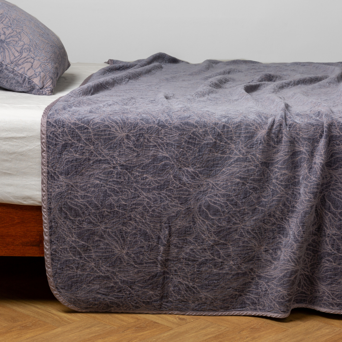 French Lavender bedspread draped over bed corner, showing botanical jacquard pattern and scalloped trim detail.