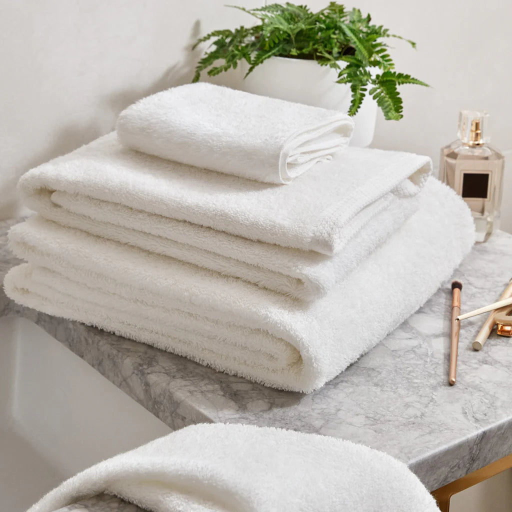 A stack of Frette Simple Border Bath Sheet Set towels sits on a marble countertop beside a potted plant, perfume bottle, and makeup brushes.