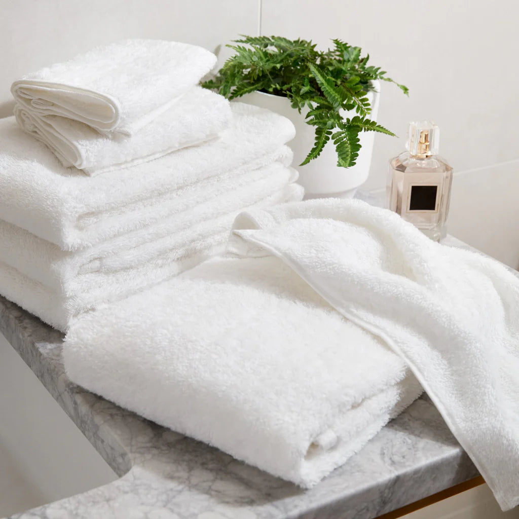 A set of Simple Border Hand Towels by Frette, crafted from 100% cotton in Turkey, rests on a marble countertop beside a potted fern and a perfume bottle in a bathroom setting.