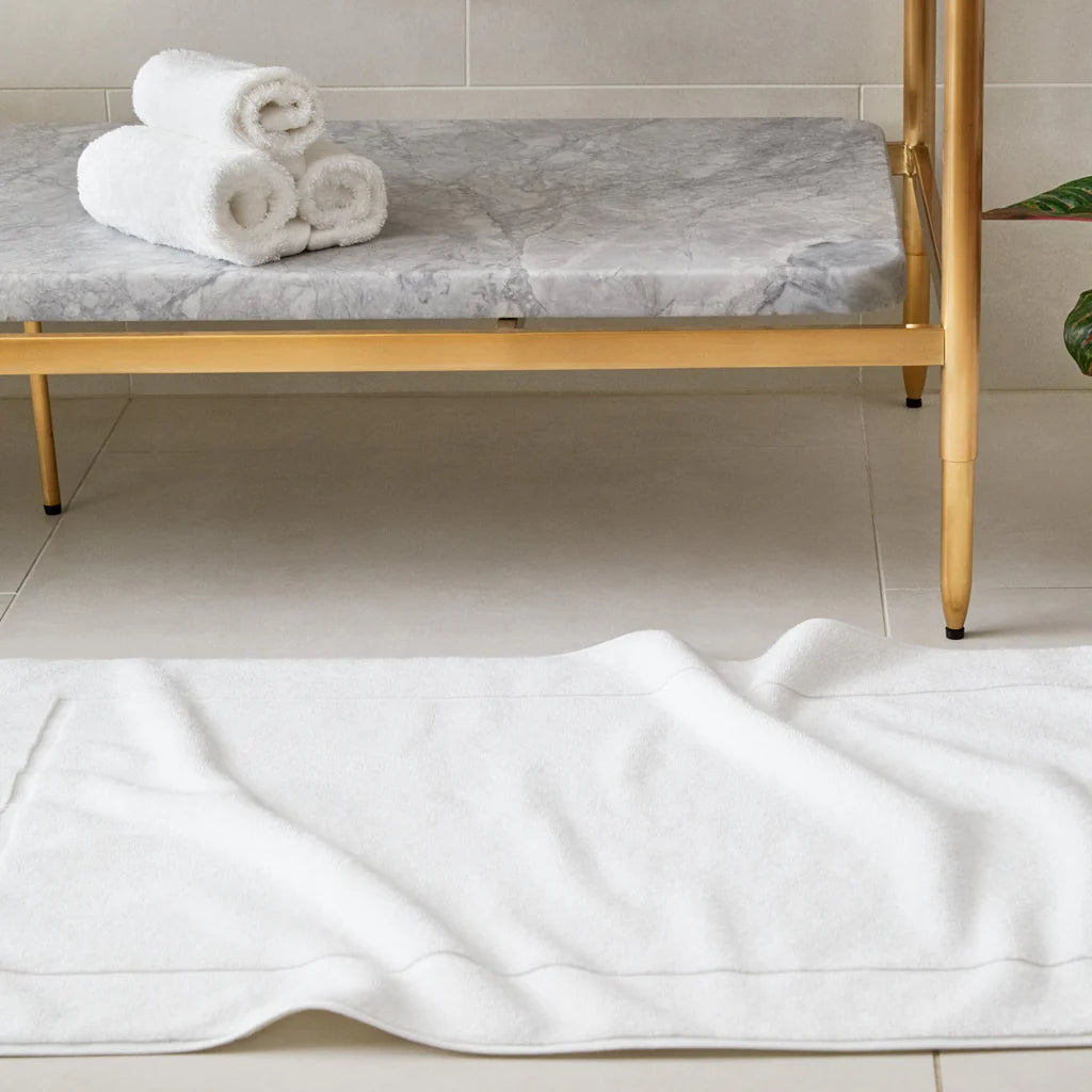 The Frette Simple Border Bath Mat is displayed on a tiled floor before a marble and gold bench adorned with three rolled luxury towels, creating an elegant and inviting atmosphere.