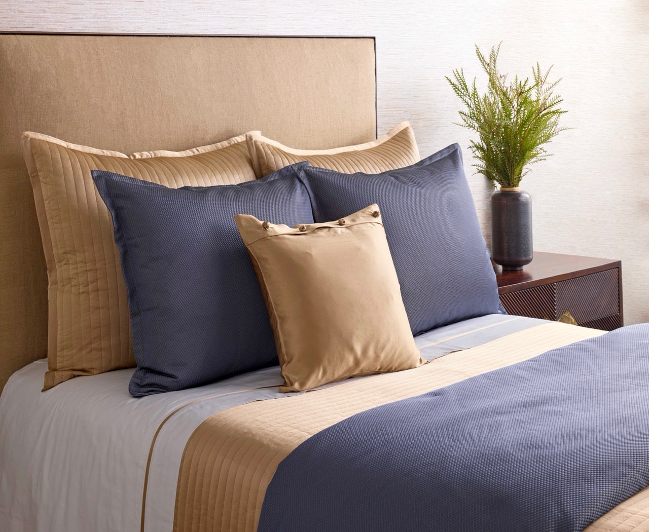 Navy and champagne bedding ensemble styled on a bed with layered pillows and a green plant accent.