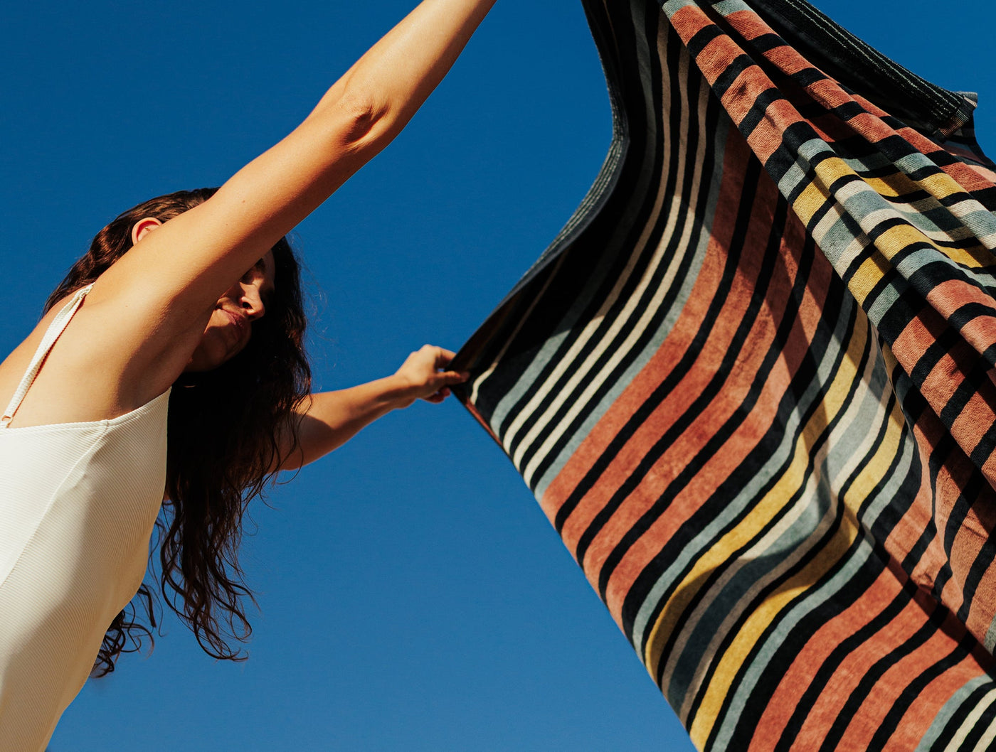 Coyuchi Dawn Stripe beach towel held aloft against vivid blue sky, multicolor stripes billowing.