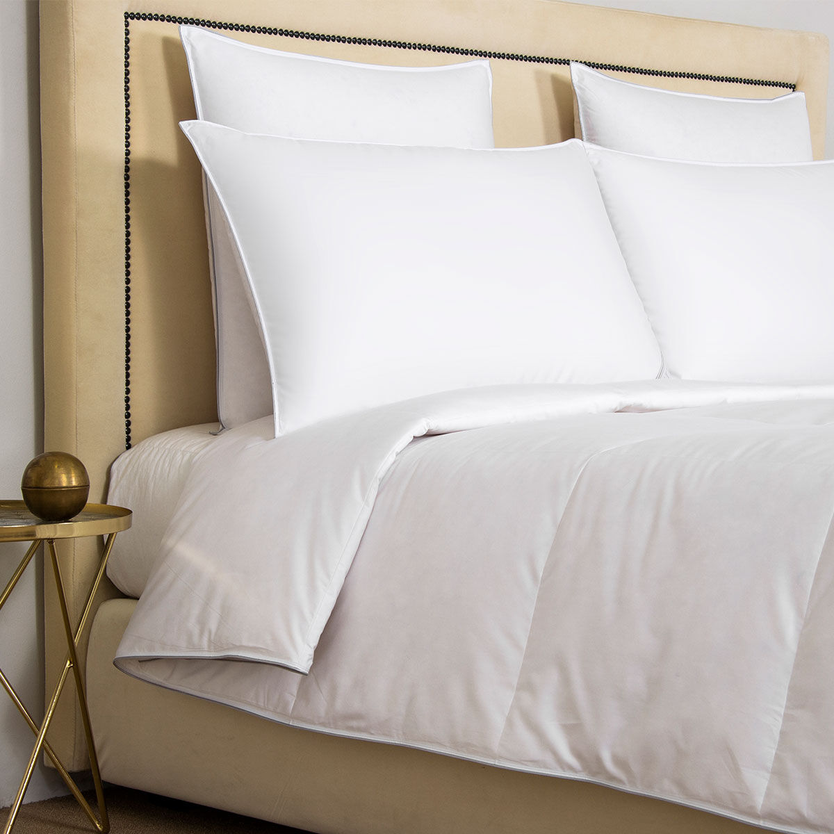 A bed is styled with the Frette Cortina Down Duvet Insert in white cotton sateen, paired with plush pillows, a beige upholstered headboard featuring nailhead trim, and a round side table topped with a gold decorative accent.