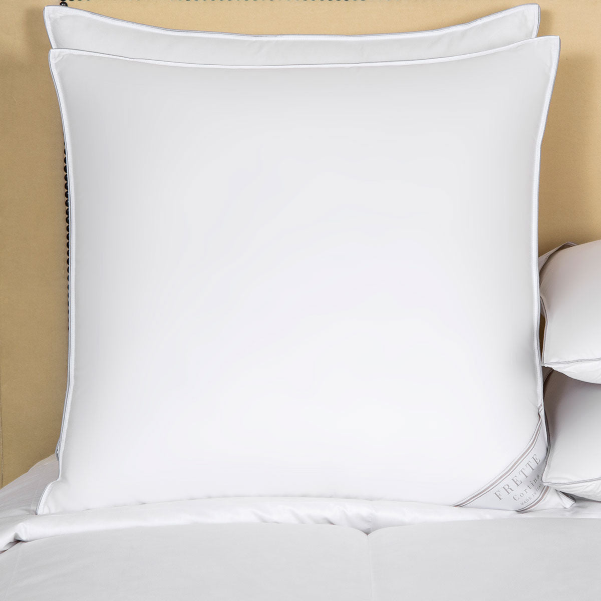 A large Frette Cortina Down Pillow with smooth cotton sateen fabric rests on a neatly made bed, part of a smaller pillow visible to the right.