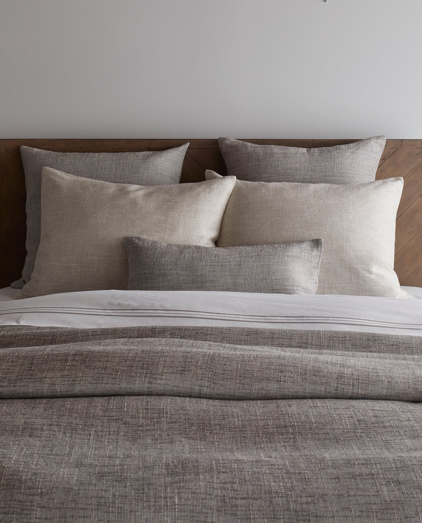 Charcoal and natural textured duvet with layered linen pillows on a wood-headboard bed.