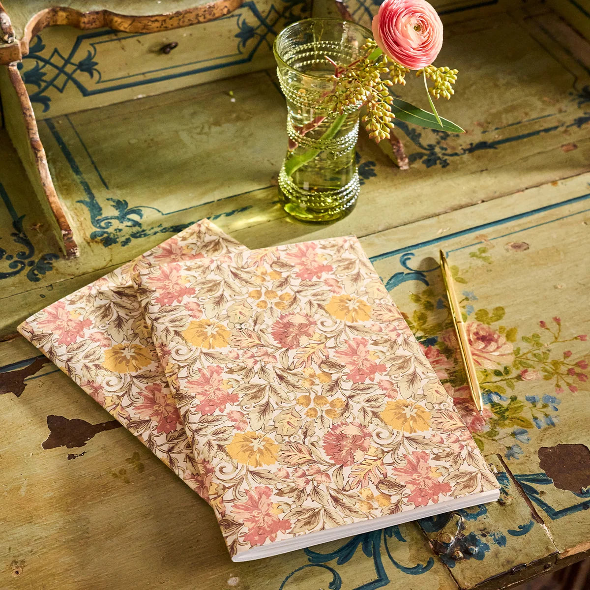 Two Juliska Willa journals on a painted desk beside a green glass with a ranunculus