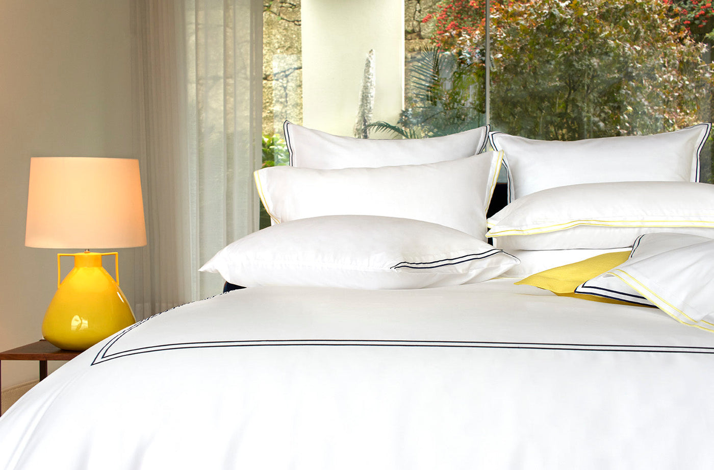 A neatly made bed features Amalia Home Collection's Sereno Sateen Boudoir luxury white linens and plush pillows, set beside a nightstand with a yellow lamp. Large windows provide views of lush greenery outside.