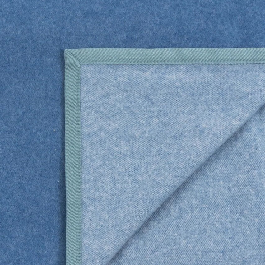 Corner detail of a sky blue wool blanket with sage green blanket-stitch trim, folded to show reverse.
