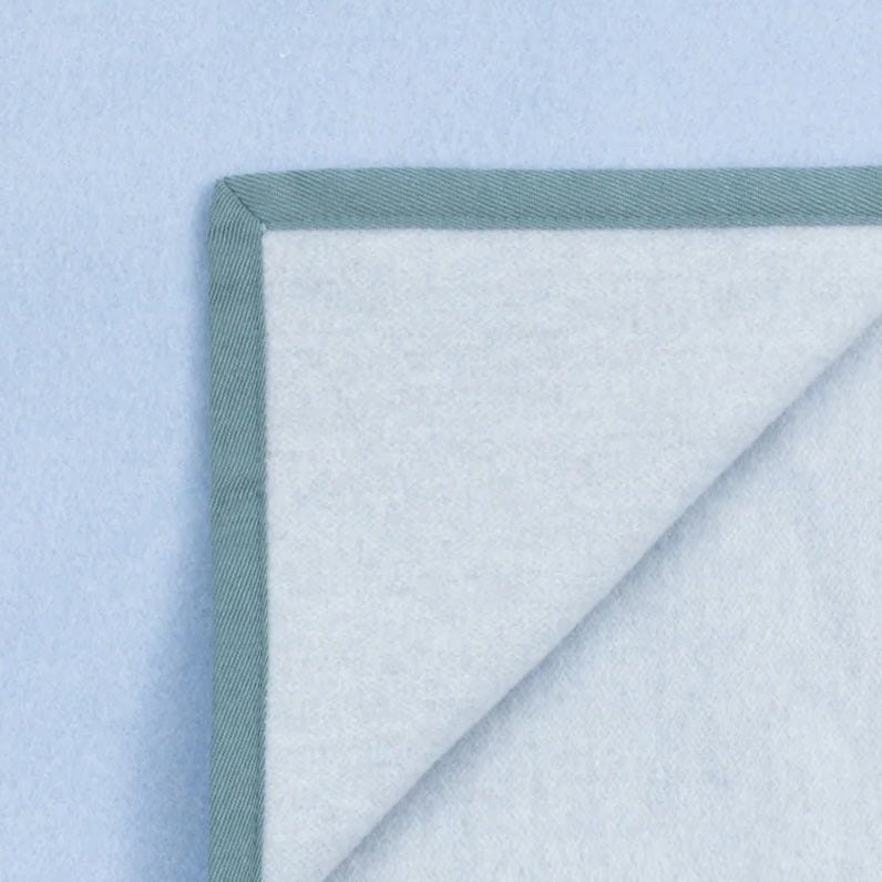 Corner fold detail showing sage green blanket binding against pale blue wool reverse.