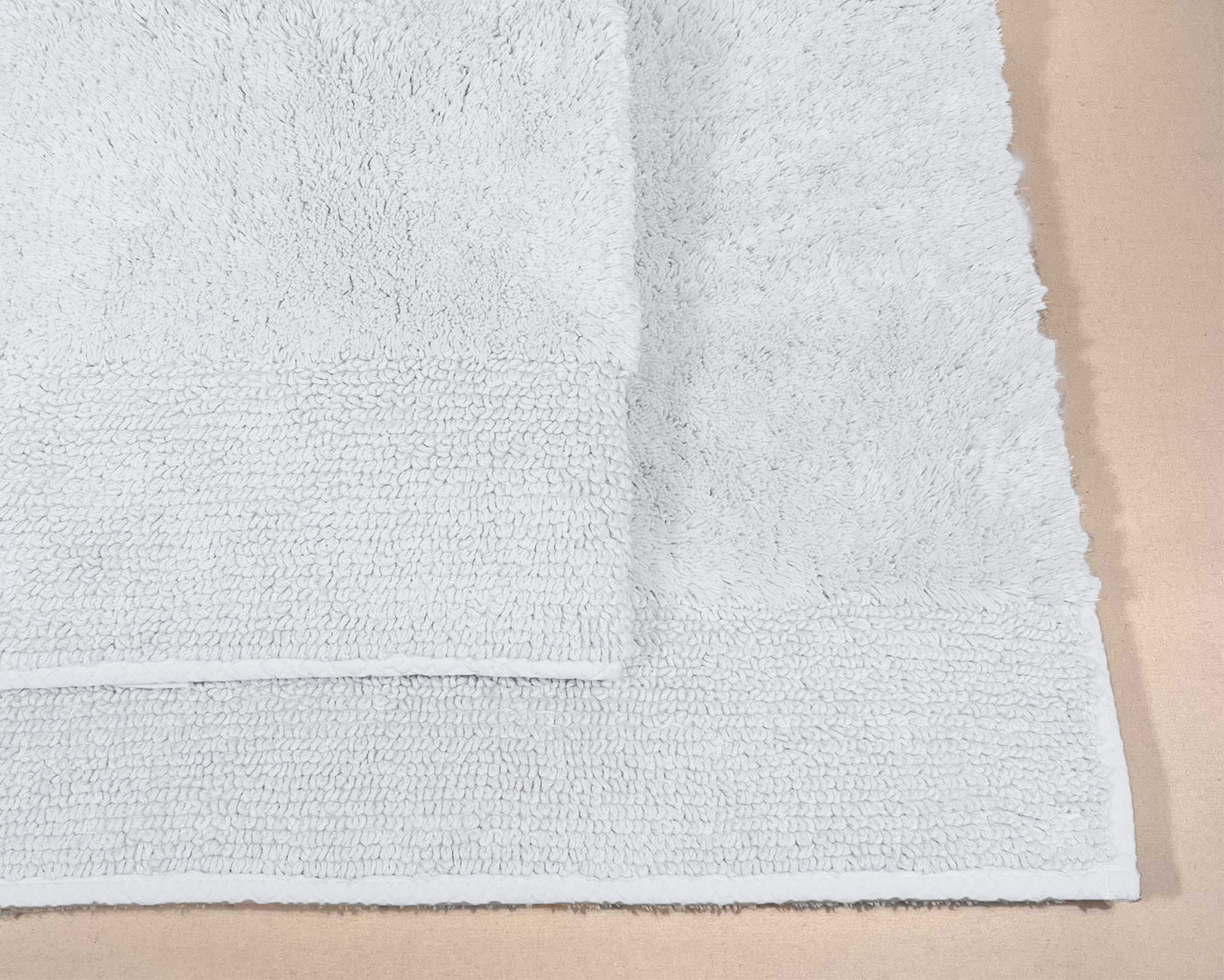 Two white bath towels, one folded over the other, are displayed on a beige surface beside the LETTO Classic Bath Rug.