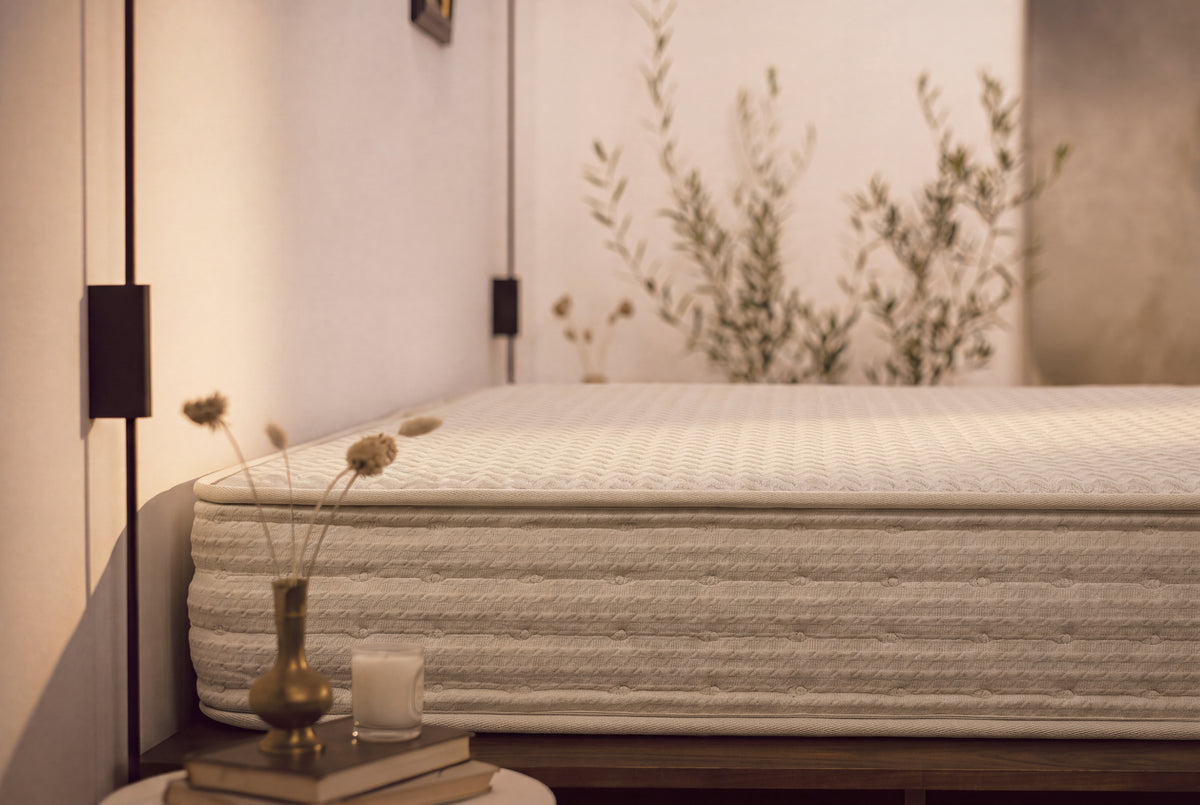 A close-up of a textured, off-white mattress in a modern, warm-toned bedroom setting with a side table, candle, and dried flowers in the foreground.