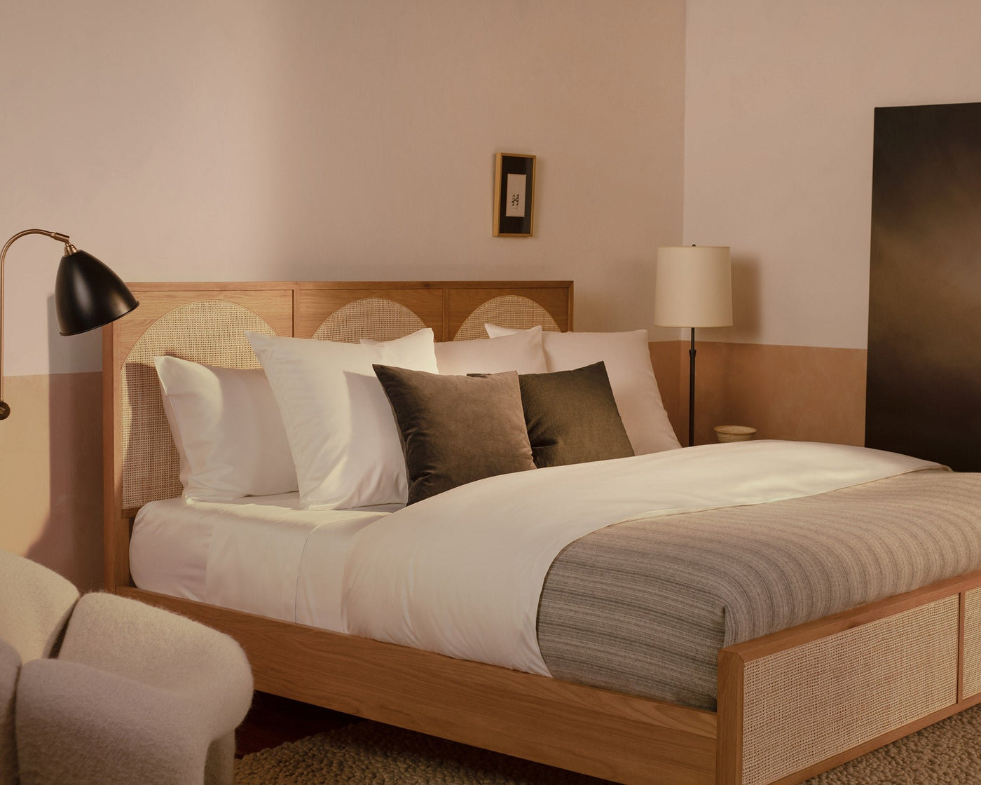 A bedroom with a contemporary wood and rattan bed made with LETTO Americano Cotton Sateen sheets, a striped gray duvet cover, and charcoal velvet throw pillows.