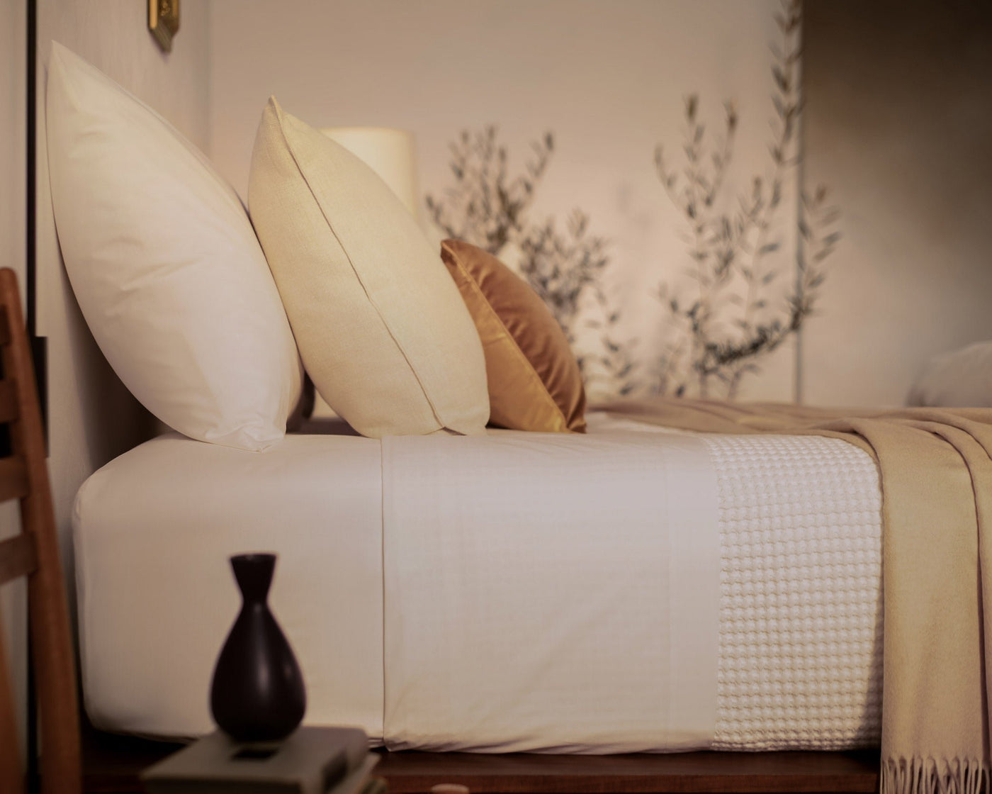 A close up picture of a bed made with LETTO White Classic Cotton Percale sheets and duvet cover, with a Nido waffleweave blanket in white and a tan velvet throw pillow.