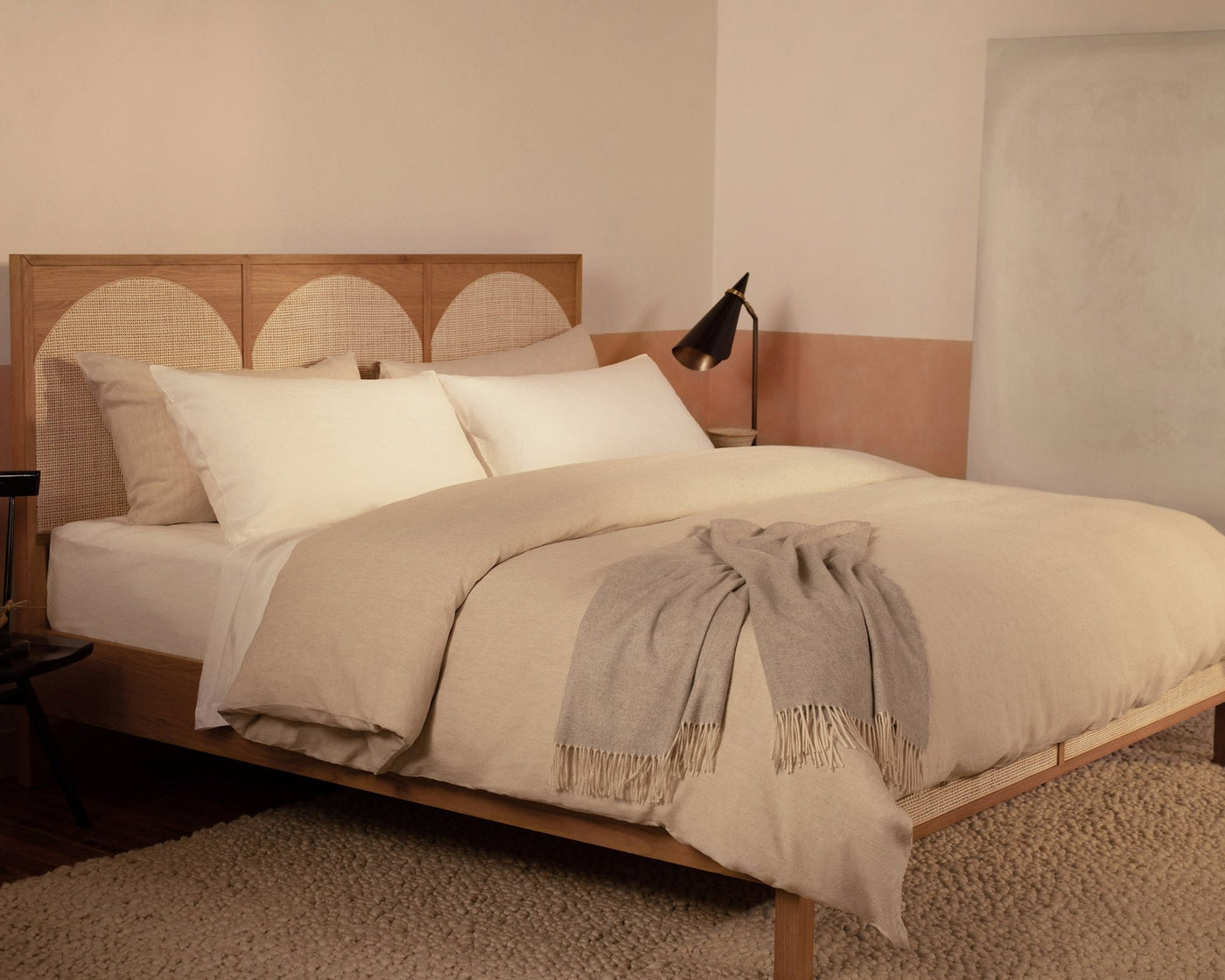 A bedroom with a contemporary wood and rattan bed made with LETTO Classic Linen sheets in white and Classic Linen Duvey in natural, with a gray Cocolla cashmere throw blanket.