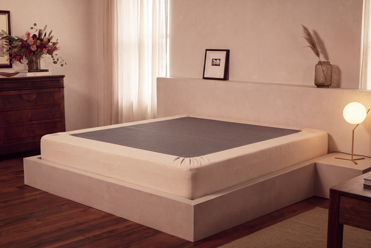 A low-profile platform bed with a cream upholstered frame and headboard, featuring a dark gray mattress topper or sheet, set in a modern, warm-toned bedroom.