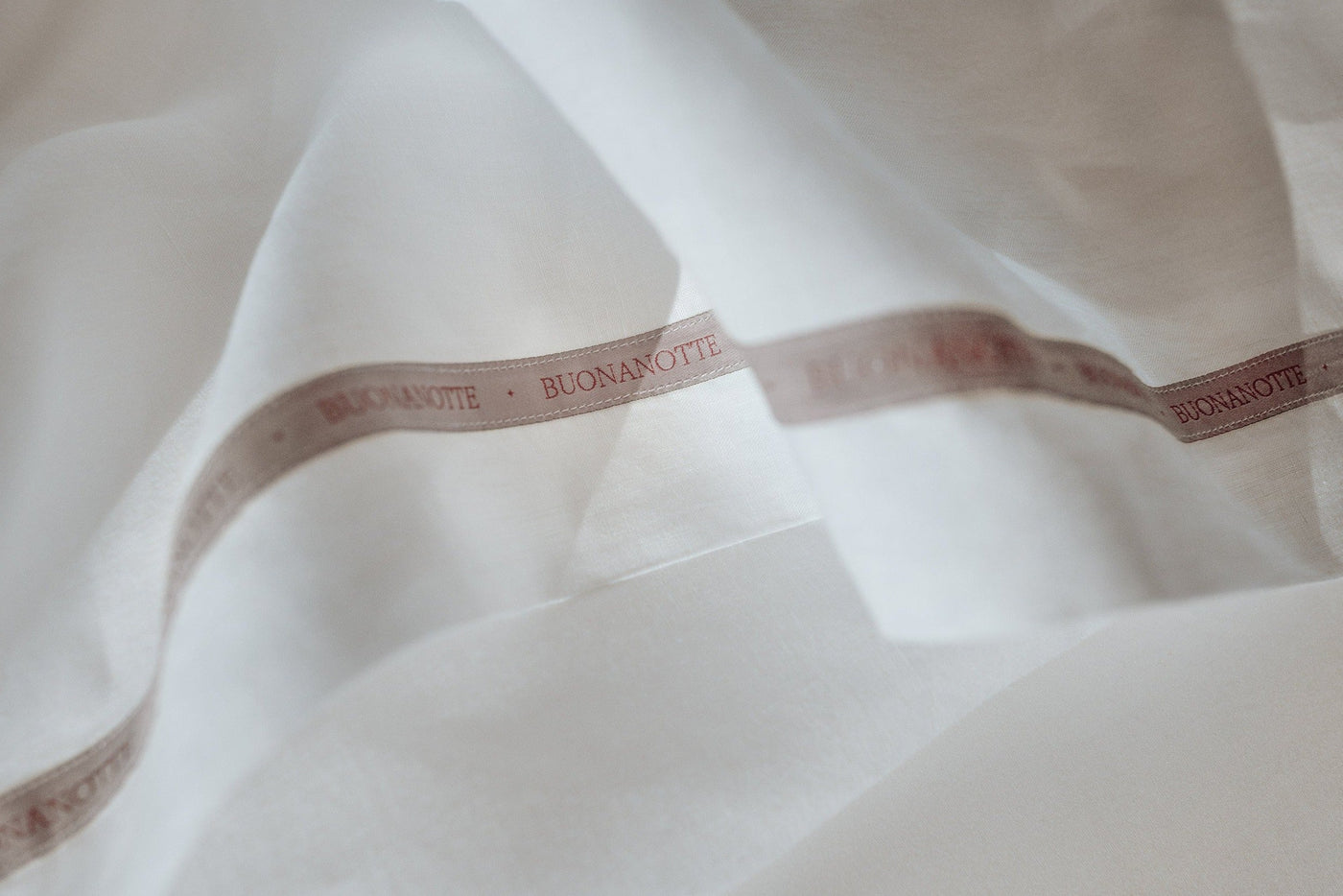 A close up image of the Buonanotte ribbon on the bottom edge of a breathable and relaxed 100% linen LETTO Lino Primo flat sheet in color white.