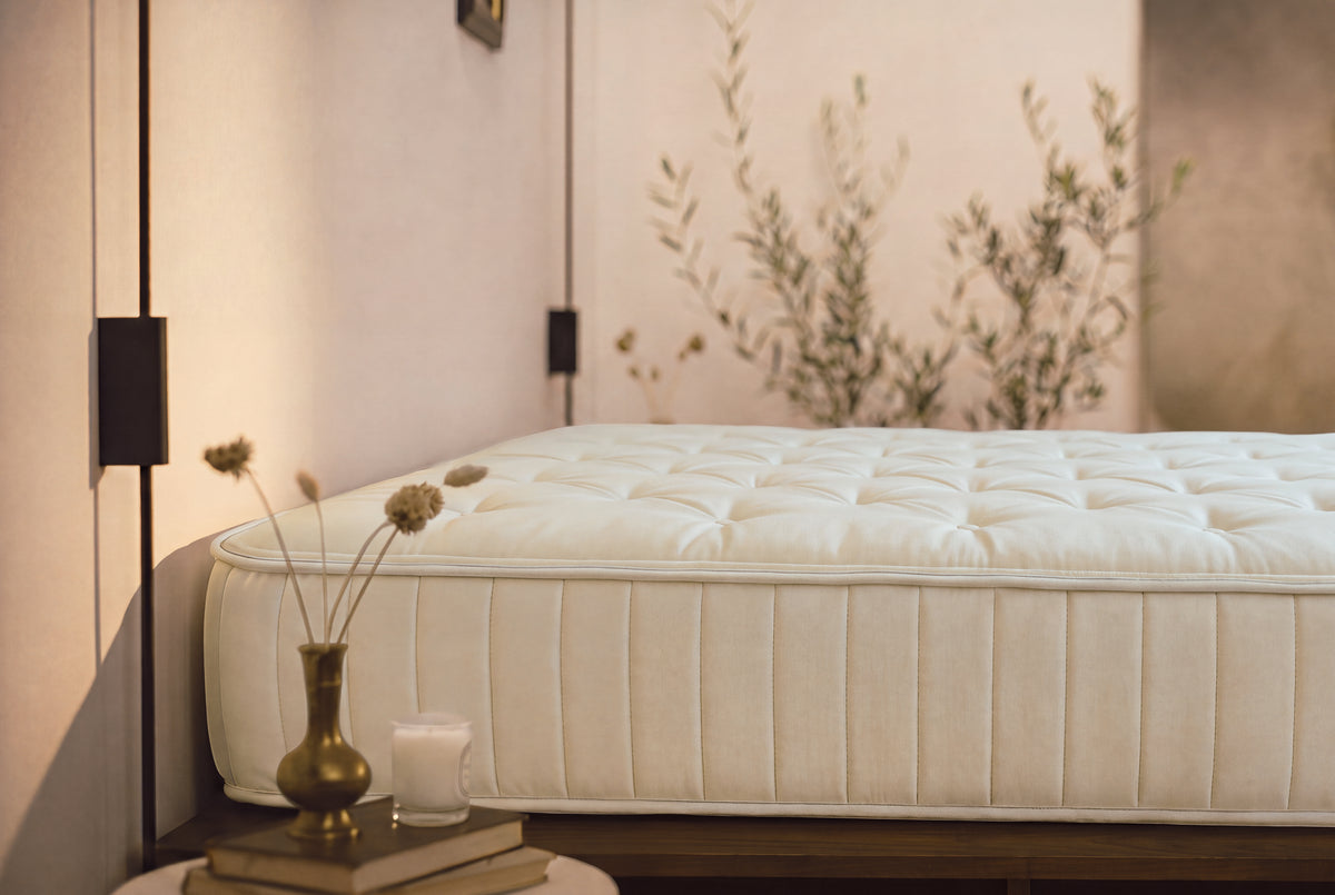A close-up of a tufted mattress corner in a softly styled bedroom setting with warm beige walls, dried stems in a brass vase, and a small candle on a bedside table.
