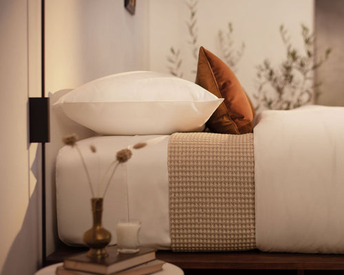 A close up picture of a bed made with LETTO White Classic Cotton Sateen sheets and duvet cover, with a Nido 100% Cotton waffleweave blanket in beige and an orange velvet throw pillow.
