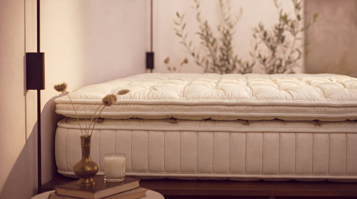 A close-up of a quilted mattress topper and mattress base in a warm, neutral-toned bedroom setting with dried flowers and a candle on a side table.