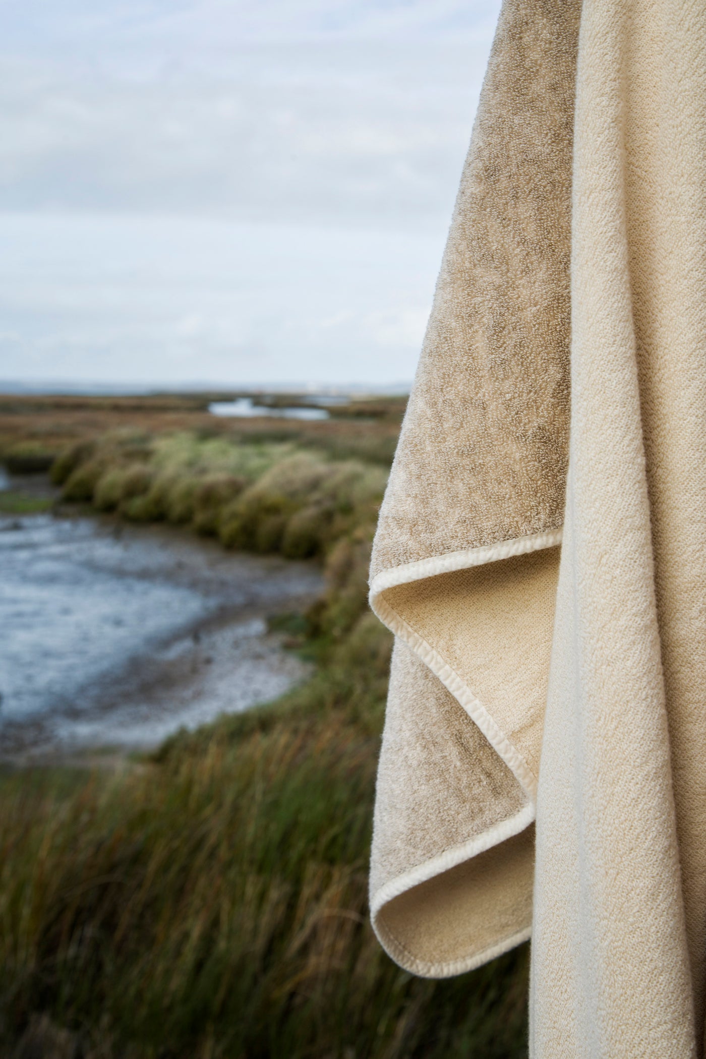 Close-up of sandy linen-toned terry cloth edge and fold against a coastal marshland backdrop.