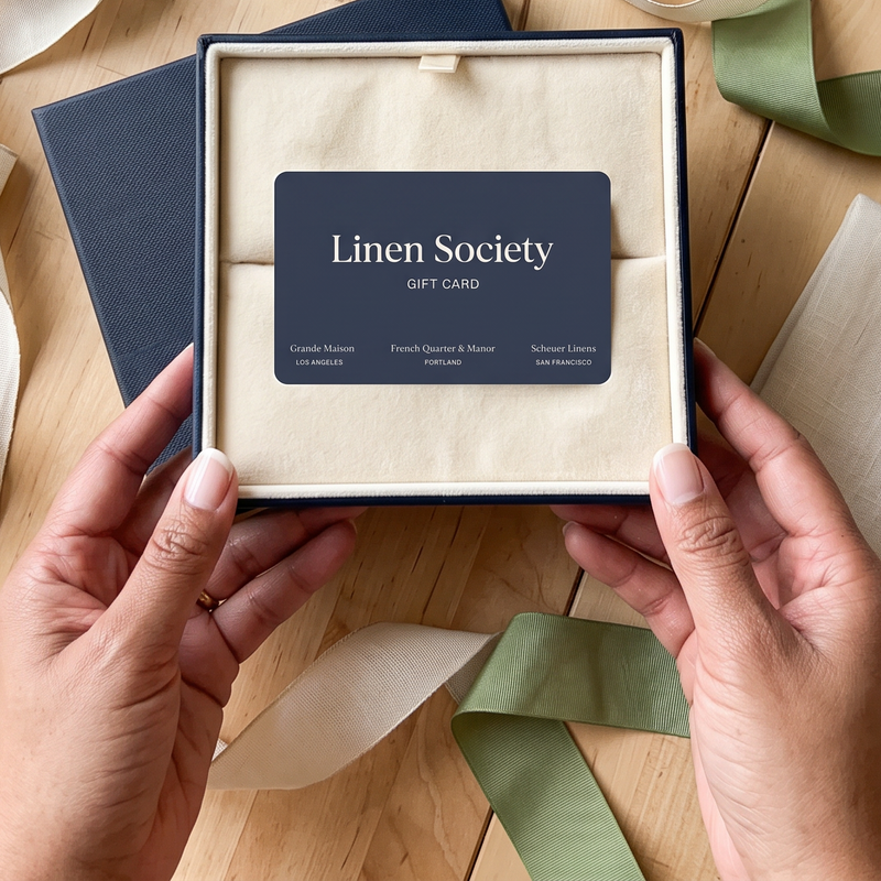 Hands holding an open navy gift box with a cream insert and a centered Linen Society gift card, surrounded by ribbons and home decor on a wooden table.