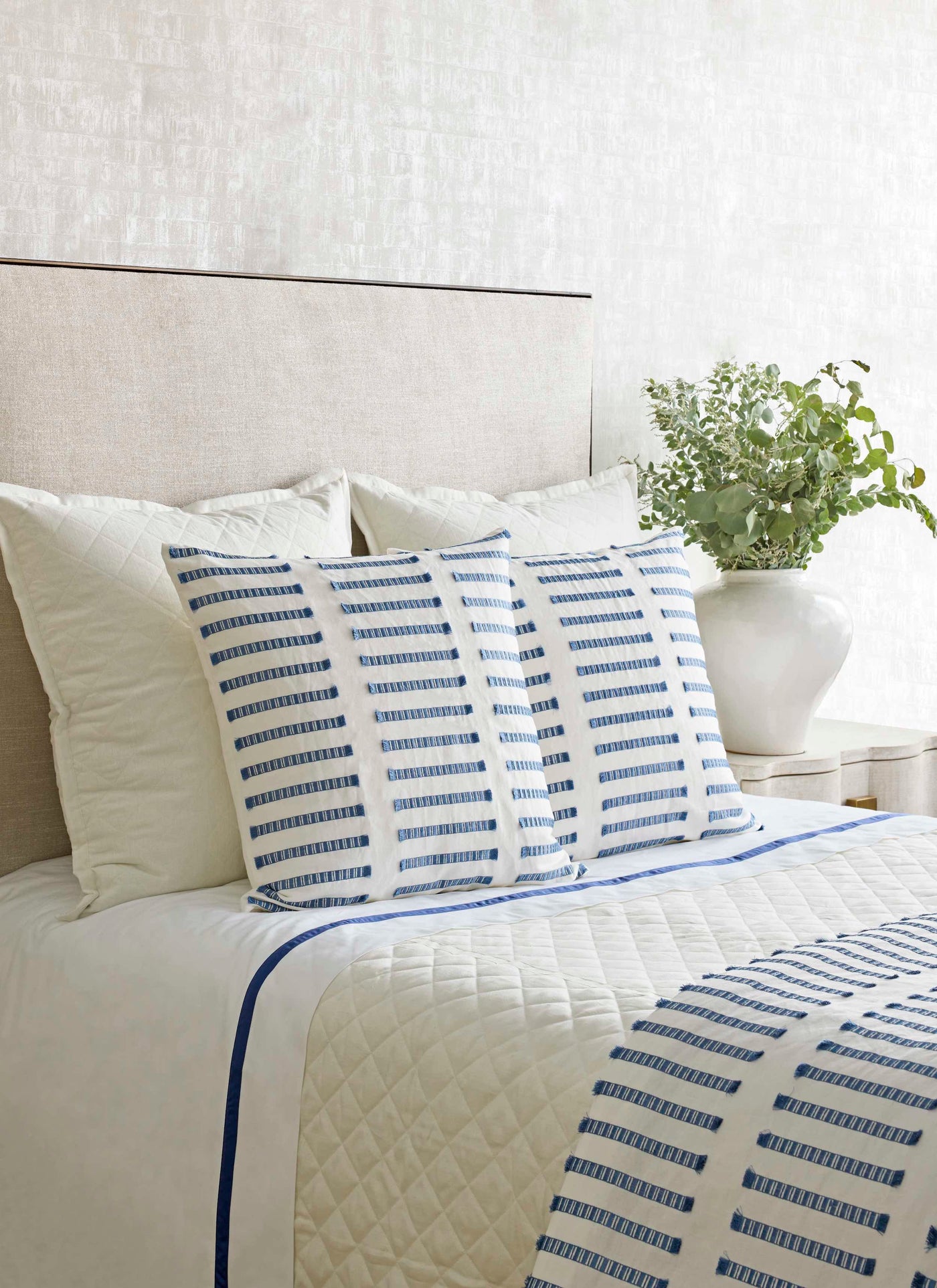 Blue and ivory striped Latera pillow styled on a made bed with quilted coverlet.