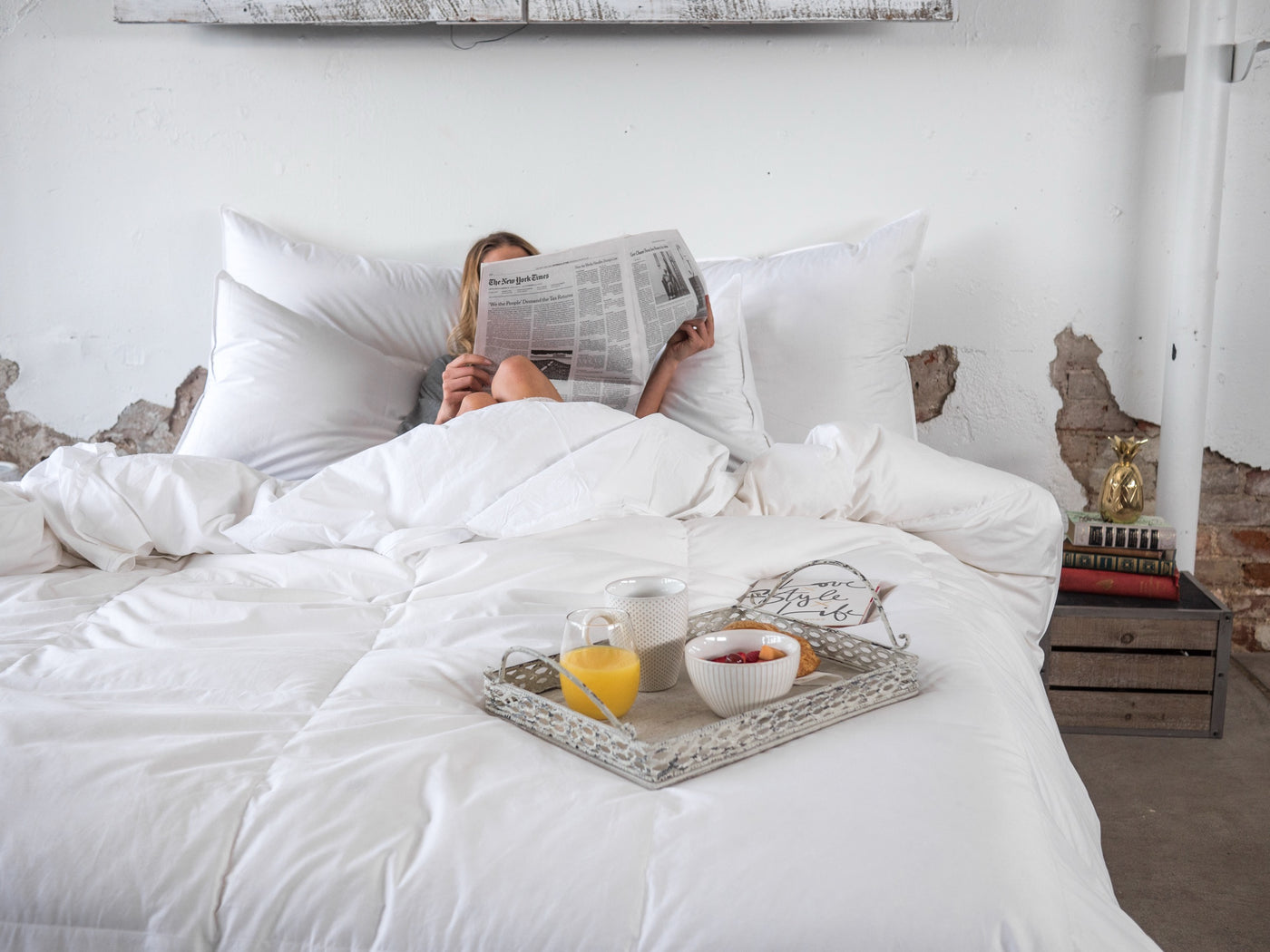 A person lies in bed reading a newspaper, covered with white bedding, with a breakfast tray on the bed in front of them. The room has a rustic white wall with exposed brick details.