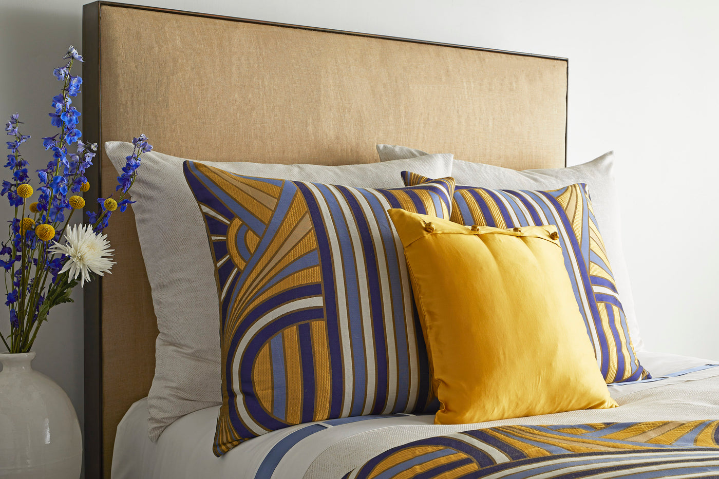 Close-up of Ann Gish Lever Du Soleil pillow in gold and blue geometric stripe pattern on styled bed.