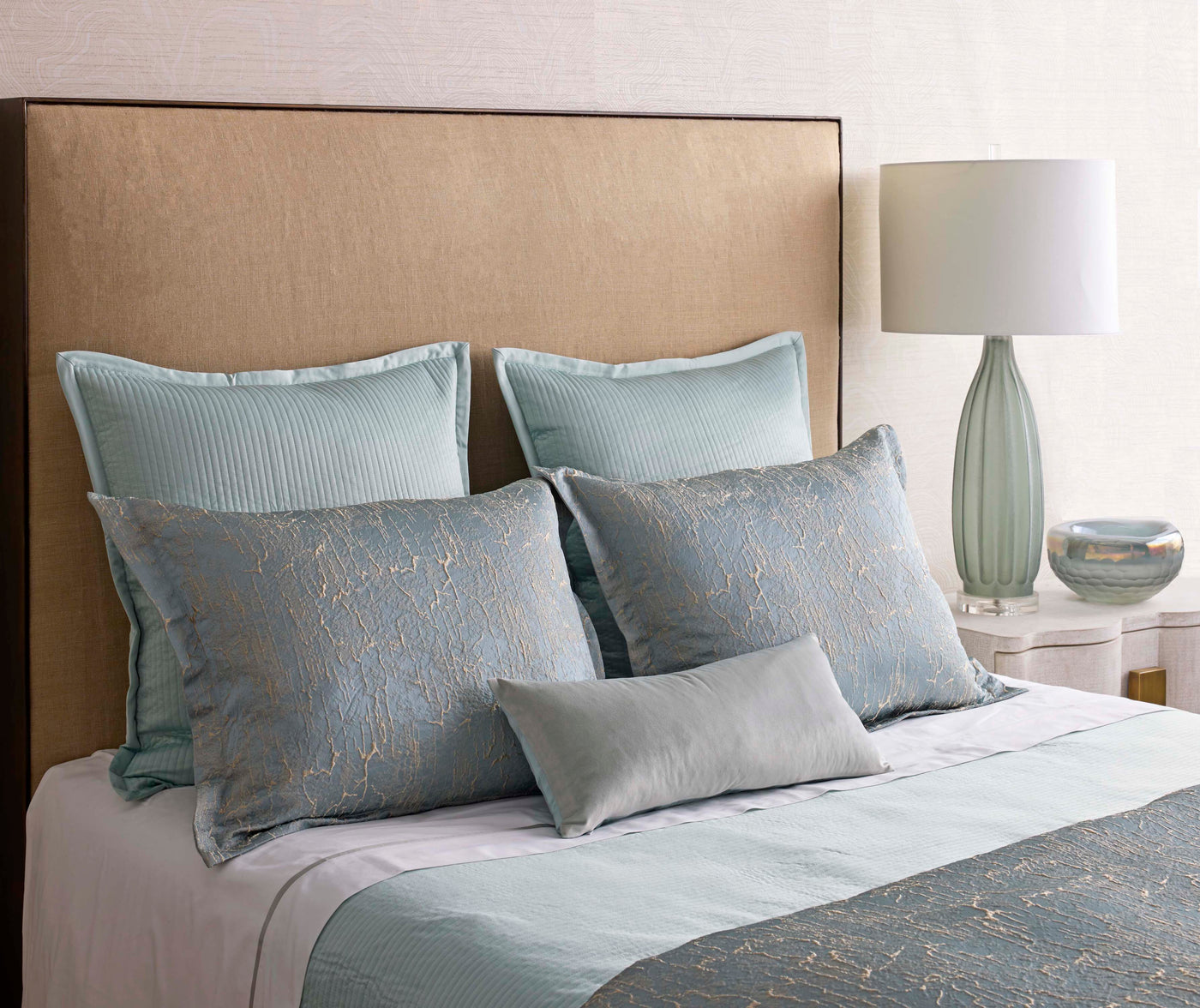 Duckegg and silver jacquard pillows layered on a styled bed with a seafoam lamp.