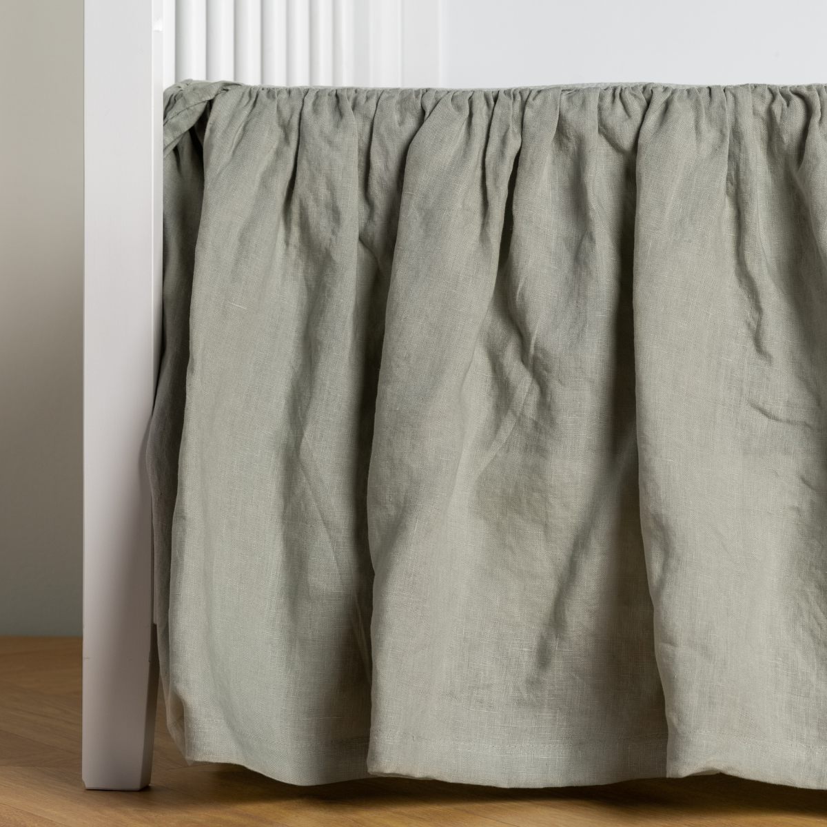 Gathered Eucalyptus linen crib skirt draping to floor, shown on white crib frame.